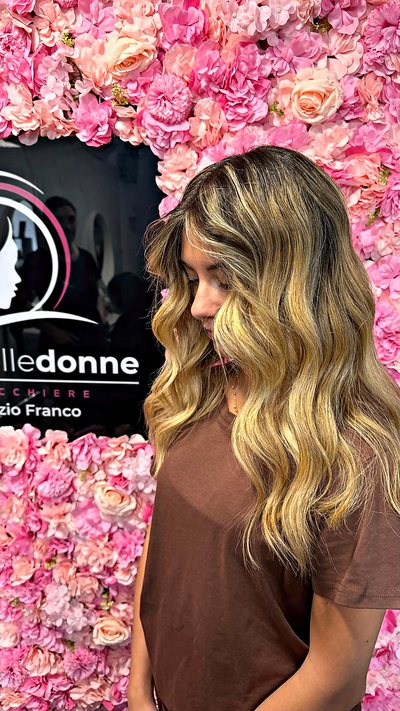 Balayage biondo su lunghezze mosse, davanti alla parete di fiori rosa del salone