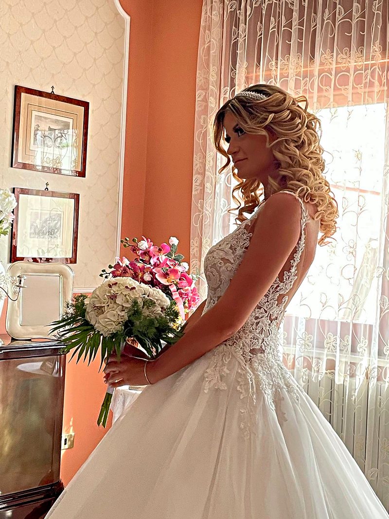 Sposa con acconciatura riccia semiraccolta, in controluce davanti alla finestra